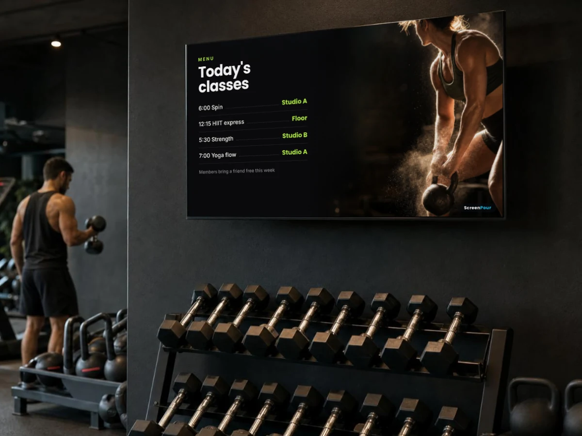 A screen on a gym wall above the weights showing todays classes next to a kettlebell photo