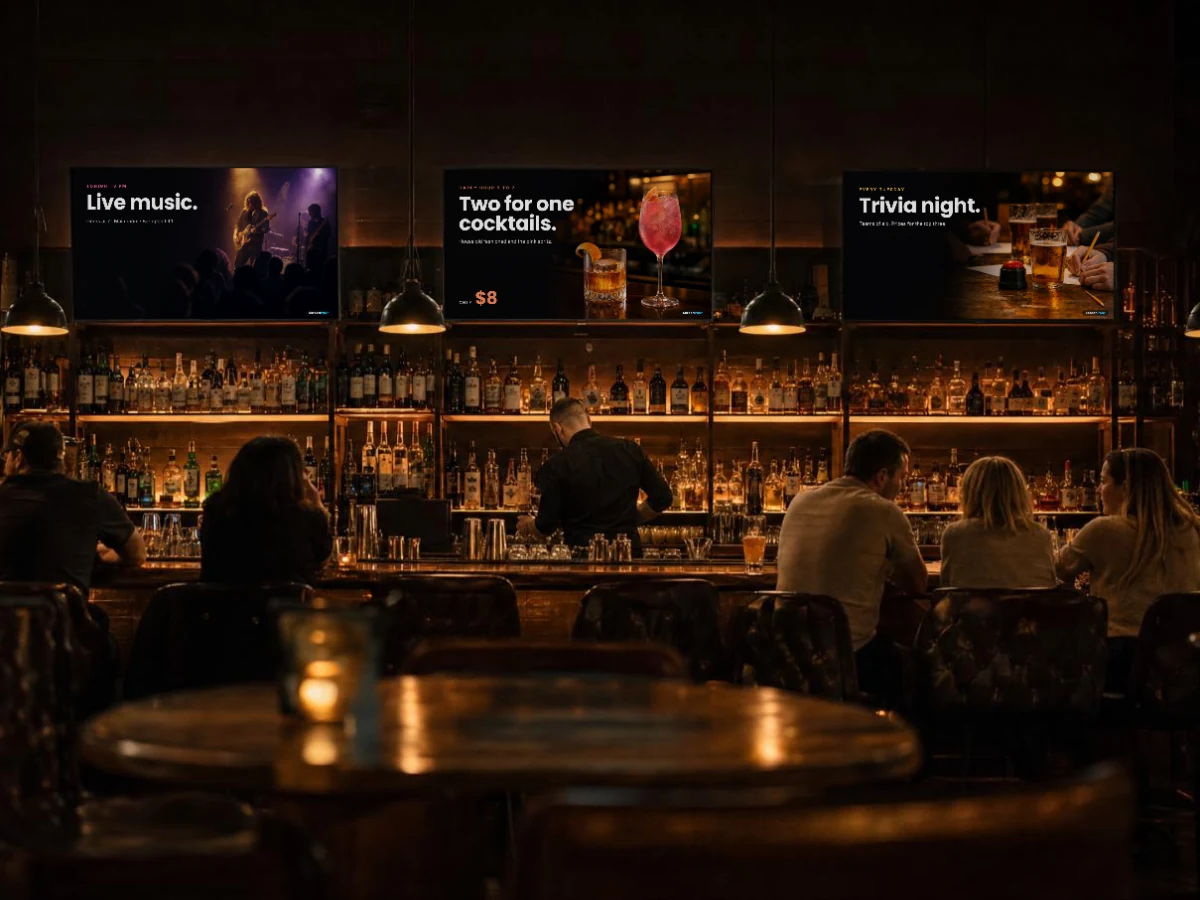Three screens above a bar at night showing live music, two for one cocktails and trivia night